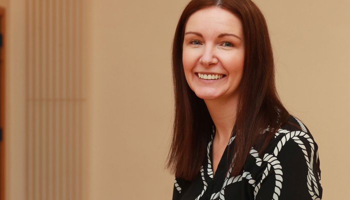 A woman with dark, shoulder-length hair wearing a black and white top is smiling as she faces the camera side-on
