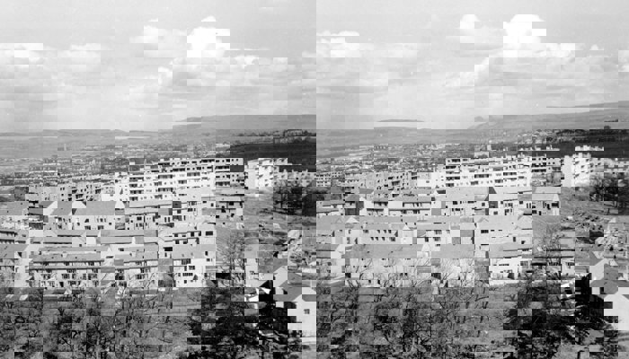 Black and white photograph of housing in Glasgow, with light coloured buildings including houses and flats with trees at the foreground and clouds overhead.