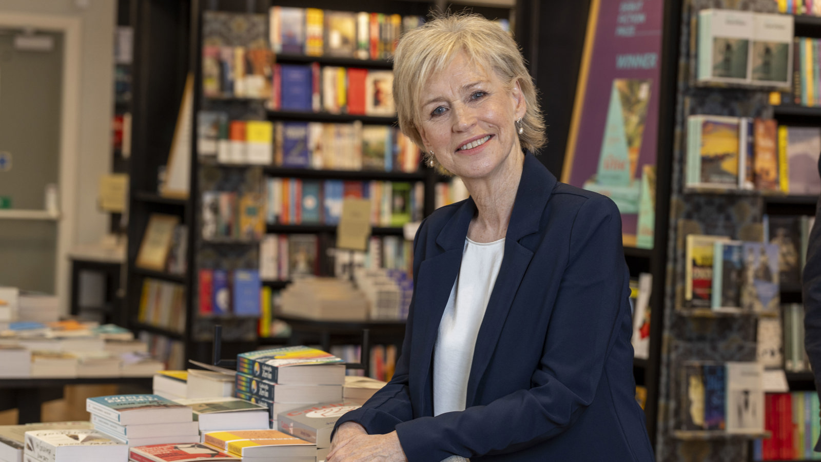 A woman with short blonde hair stands beside a table of books inside a library