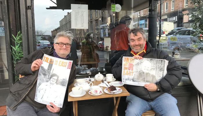 Two people sit in front of a cafe in the East End of Glasgow. The both hole portraits of people with the words Taels Fea the East written on them