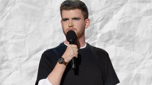 A stand up comedian holding a microphone. They have short dark hair and are wearing a black t-shirt over a white long sleeve