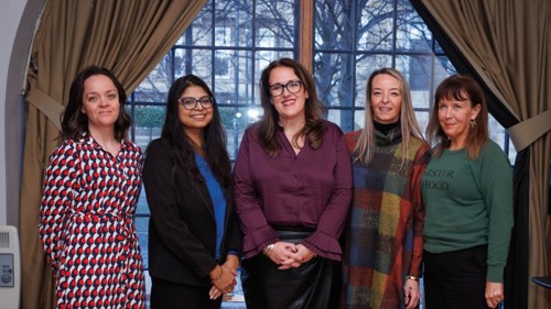 A group of five people gathered for a group photo in an arched window looking out onto bare trees and a large sandstone building behind