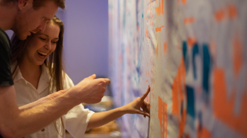 The artist show a participant the large brightly coloured hand printed banner they helped create. The banner is green and orange with geometric patterns.
