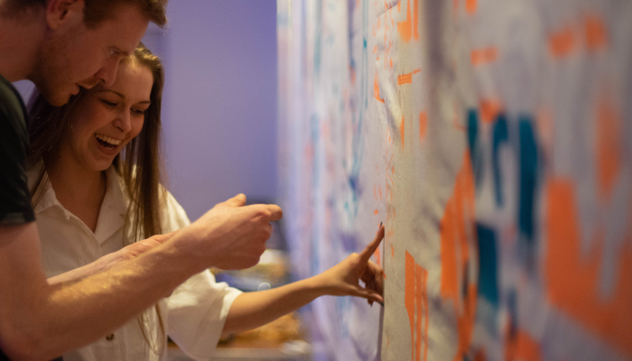 The artist show a participant the large brightly coloured hand printed banner they helped create. The banner is green and orange with geometric patterns.