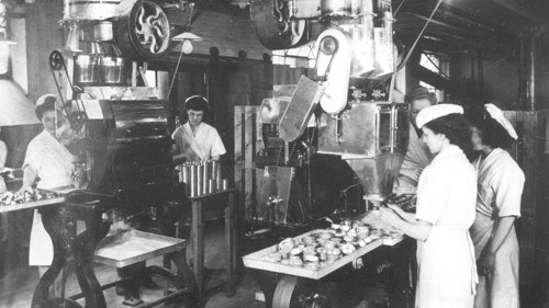 Black and white photo of people in white uniforms working on a production line with large machinery overhead.