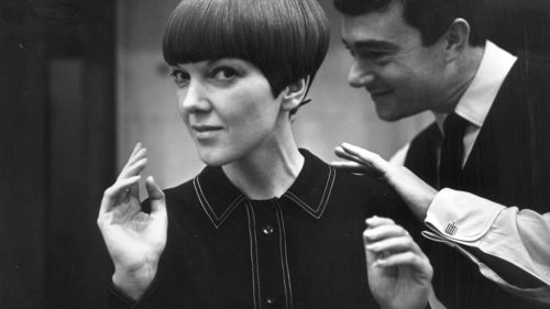 A woman with short brown hair in a buttoned top with her head turned and hand raised at shoulder height. A man is standing behind the woman with his hand on her shoulder.