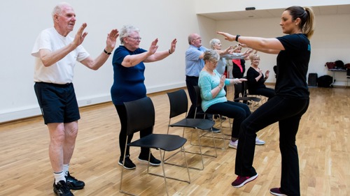 A fitness instructor taking a chair aerobics class with older participants, some are standing up while others are sitting down.