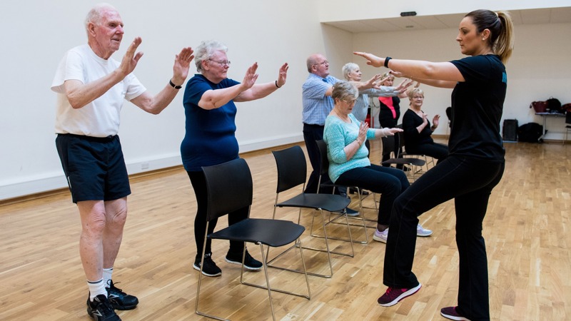 A fitness instructor taking a chair aerobics class with older participants, some are standing up while others are sitting down.