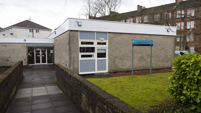 Exterior of Bailliston Library which is a flat roof building with white double doors. There is a small patch of grass outside the library and there are red sandstone tenement flats in the background.