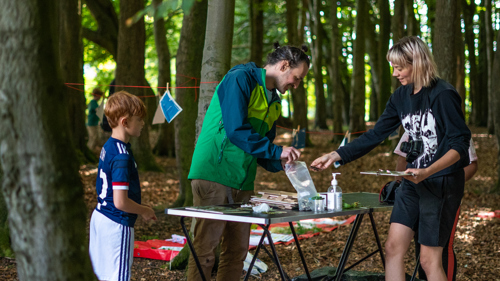 Three people stand in a wood they are smiling and looking at drawings which are displayed on a table.