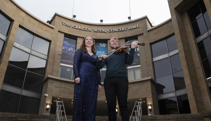 One person in a sparkly blue dress and another next to them holding a violin and bow standing on the stairs in front of Glasgow's Royal Concert Hall