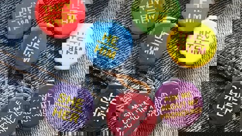 A group of colourful badges on a denim jacket showing a variety of pronouns, including She/Her, They/Them, He/ Him and Ask me about my pronouns