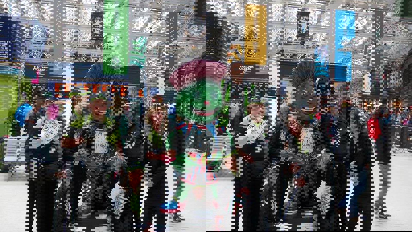 Host City volunteers in front of a Clyde the mascot statue in Glasgow Central Train Station during the Glasgow 2014 Commonwealth Games. The station is busy and has lots of Glasgow 2014 signs hanging from the roof.