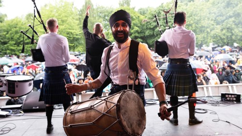 A group of musicians wearing kilts performing on a stage to an audience in the rain. Two are playing bagpipes and one a large drum