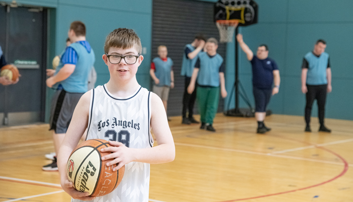 A young person stands on an indoor basketball court holding a basketball, wearing a white sports jersey. Several other players in blue bibs are gathered near the hoop in the background, preparing for or participating in a game.