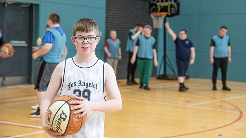 A young person stands on an indoor basketball court holding a basketball, wearing a white sports jersey. Several other players in blue bibs are gathered near the hoop in the background, preparing for or participating in a game.