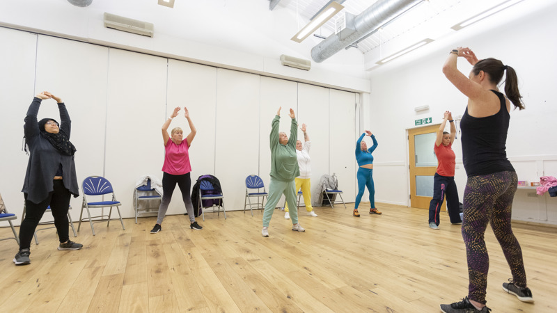 A fitness instructor standing front of six women during a class, all of them are standing up and stretching with their hands raised over their heads.