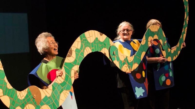 Two elderly people perform on stage with a giant snake cut out. 