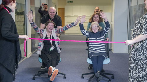 A group of people in an office cheering on two colleagues being pushed in office chairs towards a finish ribbon. The people in the chairs have their arms raised