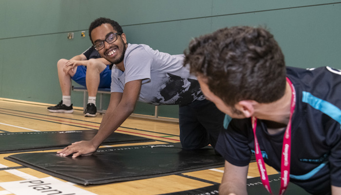 Two men are smiling and exercising on mats on the floor of a sports hall.