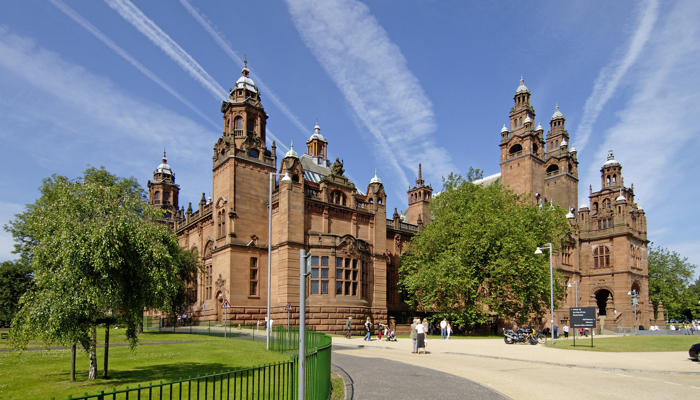 A red sandstone building beneath a blue sky is surrounded by green trees