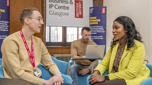 Two people sitting in armchairs having a conversation in a library. There is a person working on a laptop behind them.