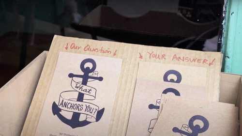 Specially made postcards sit in a box. They show an anchor with a banner wrapped around it reading "what anchors you?"