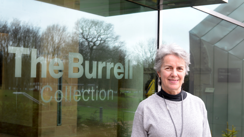 A person standing outside the Burrell collection. They have grey hair and a grey jumper with a lanyard around their neck. They are smiling at the camera.