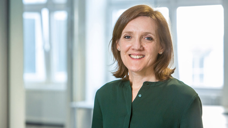 A person with shoulder length brunette hair, wearing a green top, smiling in front of some large windows in an open plan office space
