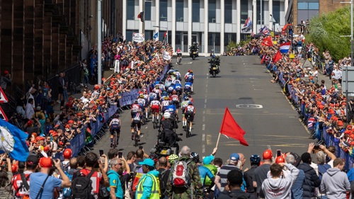 A cycling road race in a city centre crowded with spectators. Cyclists in colourful kit are racing up a steep hill on a clear day