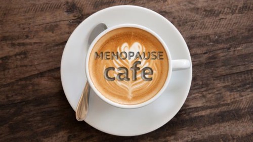 An overhead shot of a cup of coffee on a table with the words Menopause Cafe written in the foam on top of the coffee
