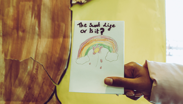A hand holds a drawing with a rainbow with the questions "it's a sweet life, or is it"
