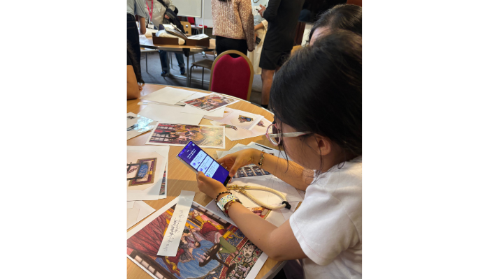 A person sits at a table, which is covered in papers. They are holding a mobile phone.