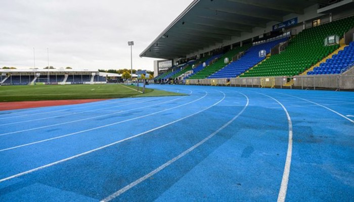 Section of a blue outdoor running track in a stadium. There are large tiered seating areas to the sides and a grassy area in the middle of the track.