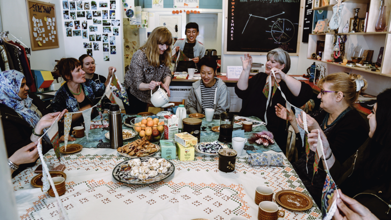 Many community members sit around a large table laughing and sharing stories. There are cakes, tea and treats that are being served to everyone. 