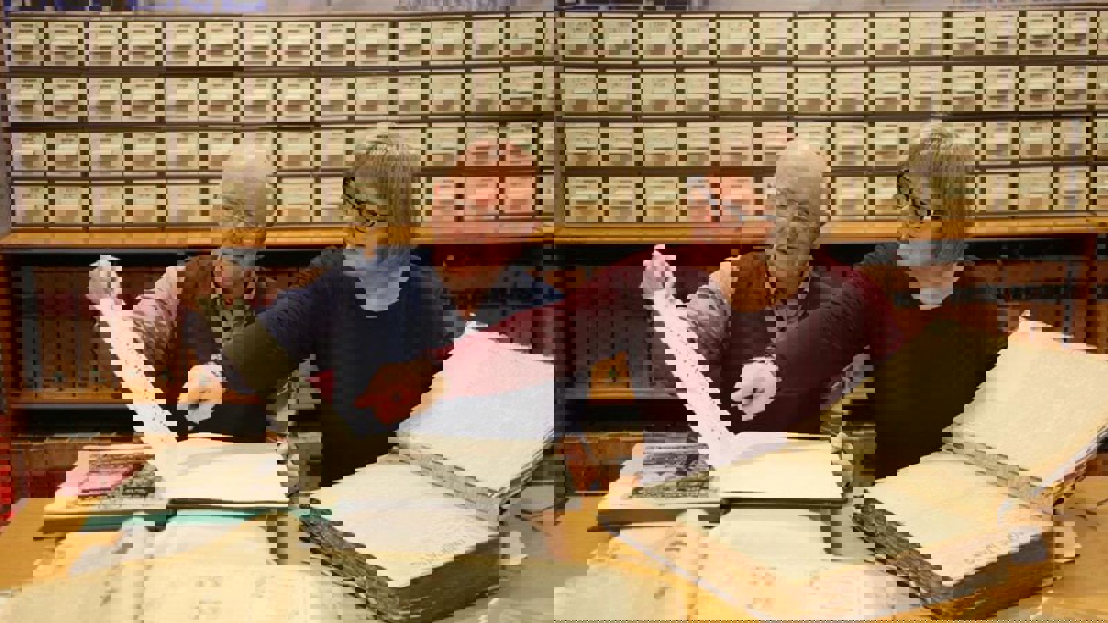 Two people sitting at a desk with two large old books in front of them with filing cabinets behind them in the background