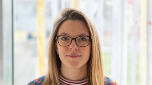 A person with glasses and long blonde hair stands in front of a window smiling. 
