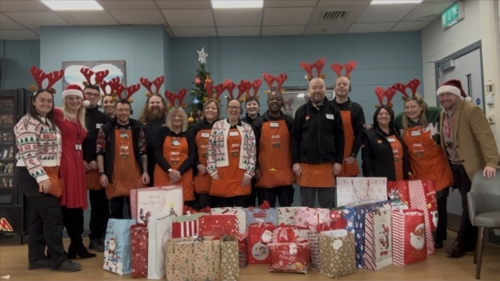 A large group of people dressed in orange aprons and reindeer antlers are standing behind a large pile of wrapped Christmas gifts in a store breakroom