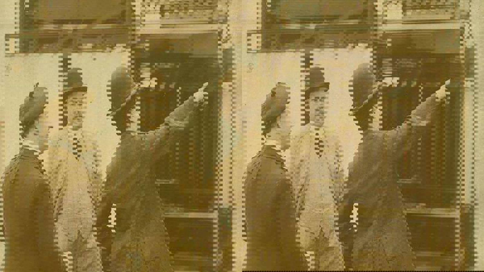 A black and white photograph of three people, including Harry Lauder and Charlie Chaplin in a warehouse, all wearing hats and one pointing to something in the distance.