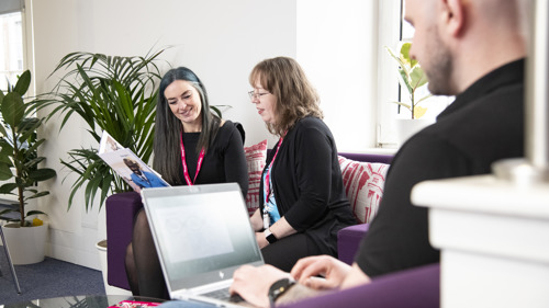 Three people working on sofas in an open plan office. One is working on a laptop, the other two are looking at a shared folder