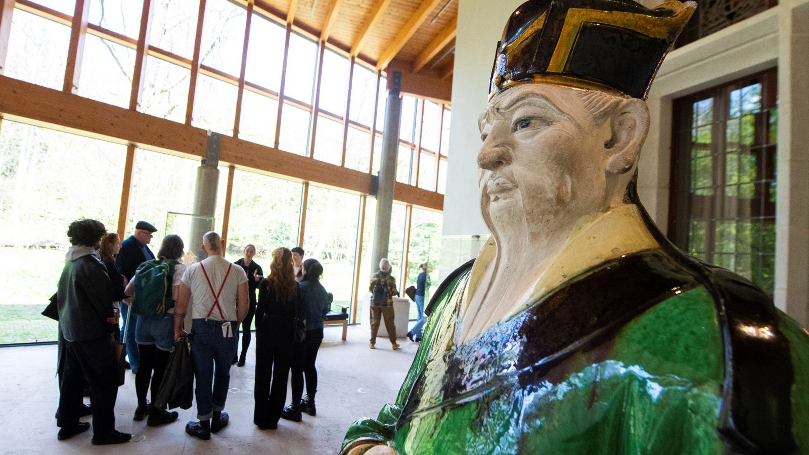 A group of people taking part in a guided tour at a museum. They are in a room with large floor to ceiling windows looking out onto a forested area and there is a large ceramic figure in the foreground