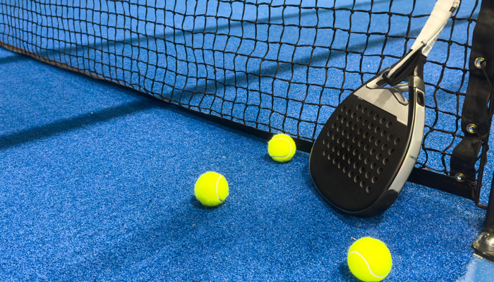 A padel racquet is propped up against a net on a padel court. Three padel balls are beside the racquet.