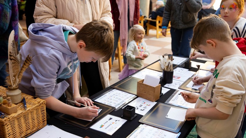 Children and adults are gathered around a large table, they are filling in cards with pencils