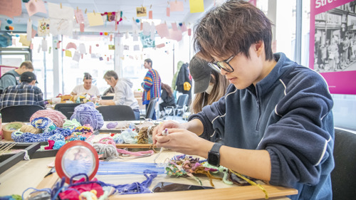 A picture of a person weaving on a loom in a busy and colourful room of people taking part in craft activities.