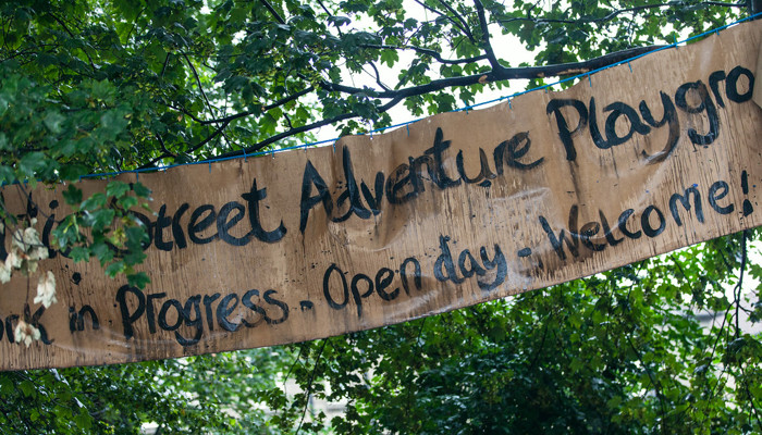 A sign hanging between trees and leaves which says Baltic Street Adventure Playground.