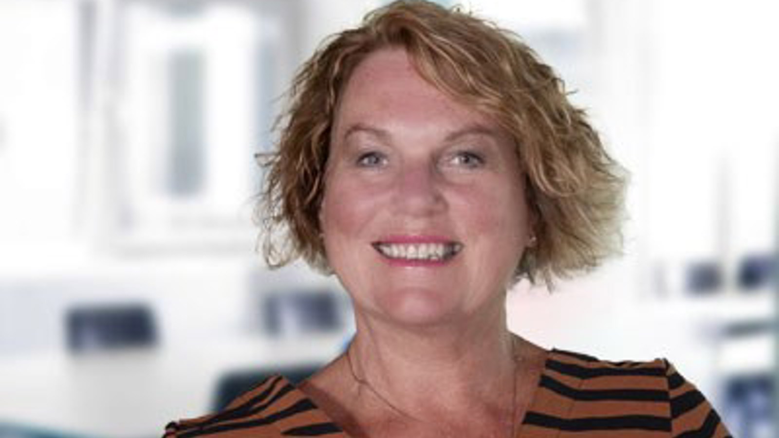 A headshot of Jan Buchanan, a person with blonde wavy hair wearing a brown and black striped top