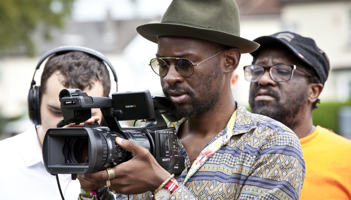 A person with sunglasses and a hat films on a professional camera.