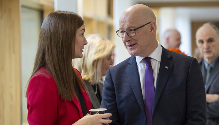 A woman in a bright red blazer speaks with a bald man wearing glasses and a dark suit with a purple tie in a modern indoor setting. The woman holds a paper coffee cup as they converse. Several other people stand in the blurred background of the hallway.