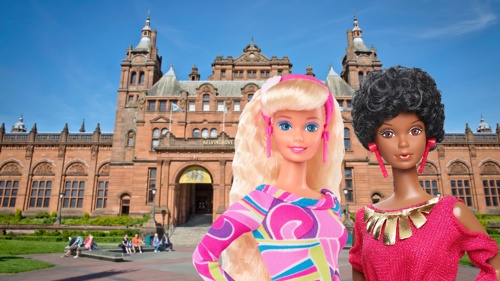 Two fashion dolls are posed in front of the frontage of Kelvingrove Art Gallery and Museum, a large, ornate, red sandstone building on a sunny day. The doll on the left has long blonde hair and wears a bright patterned pink, purple, and green dress with pink accessories. The doll on the right has short curly black hair and wears a red dress with gold details. Behind them, the museum’s red sandstone frontage, towers, and arched entrance are visible, with people sitting and walking in the foreground. 