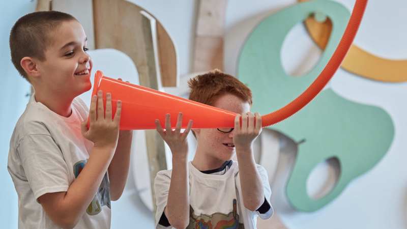 Two young boys are playing with an orange object in a bright airy room with irregular shapes in the background